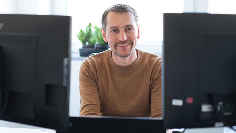ConnectedCare Projektmanager lächelnd zwischen zwei Monitoren