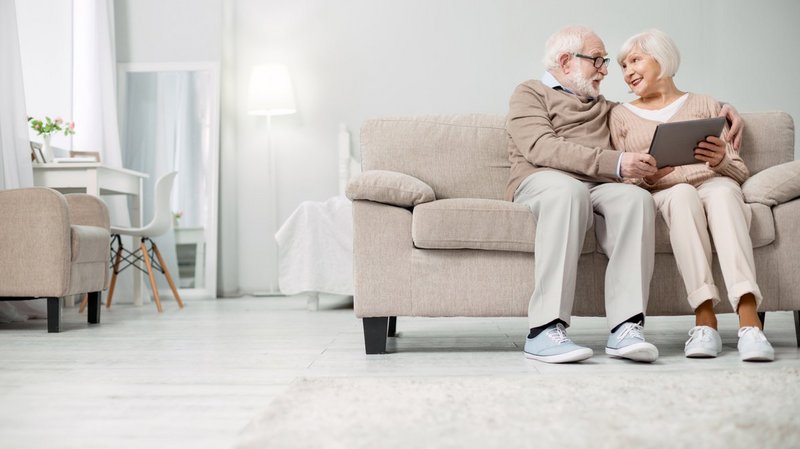 ConnectedCare Third Party Partner | GOLDENSUMMER.TV, Senioren-Ehepaar sitzt Arm in Arm auf Sofa im Wohnzimmer mit Tablet