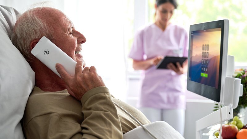ConnectedCare Patientenkommunikation | Patient liegt im Bett und telefoniert mit ConnectedCare Phone, Pflegerin im Hintergrund