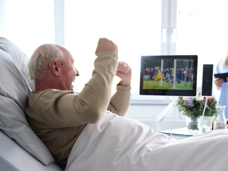 Patientenentertainment: Älterer Patient sitzt jubelnd im Krankenhausbett und schaut Fußball auf Bedside Terminal