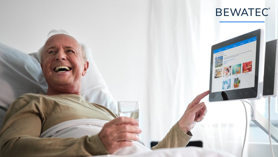 Älterer Patient im Krankenhausbett bedient lachend das ConnectedCare Tablet und hält ein Glas Wasser in der Hand