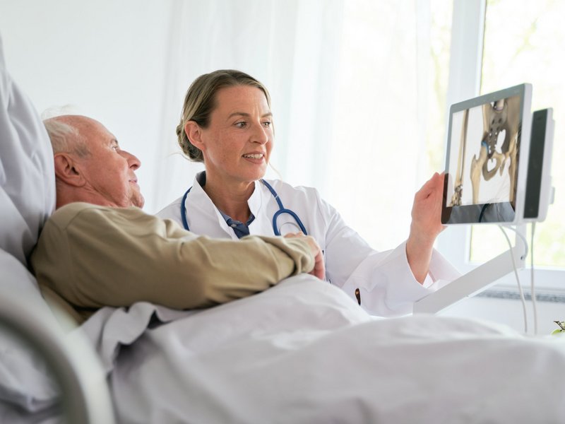 Patienten informieren: Ärztin zeigt Patient im Krankenhausbett Röntgenaufnahmen auf Bedside Terminal