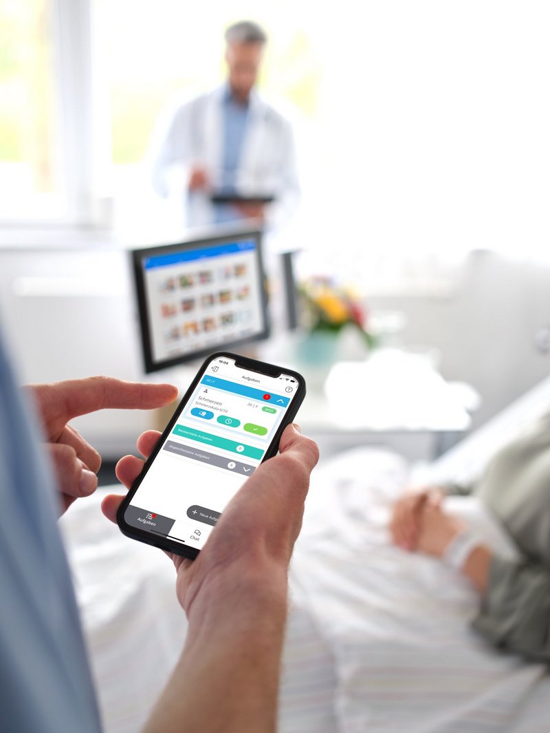 Digitale Patientenkommunikation: Pfleger steht am Patientenbett einer jungen Frau und bedient Smartphone mit dem Digitalen Serviceruf.