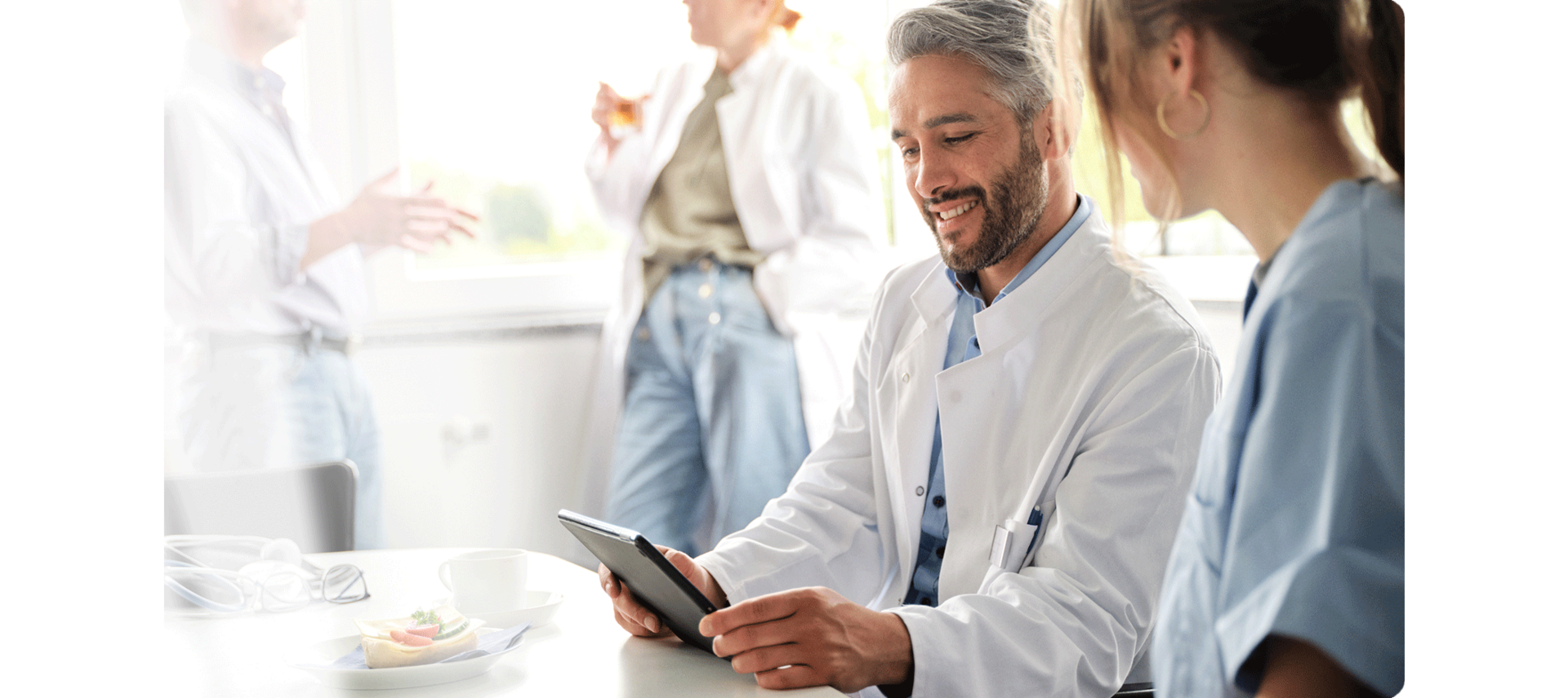 ConnectedCare Krankenhaus digitalisieren | Arzt sitzt mit Pflegerin am Tisch im Pausenraum und schaut auf Tablet, im Hintergrund unterhalten sich zwei Ärzt:innen