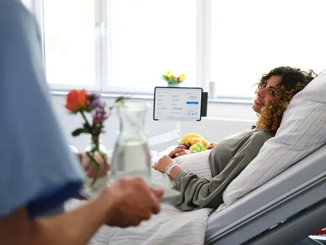 Pfleger bringt einer Patientin eine Karaffe Wasser und Blumen zum Bett.